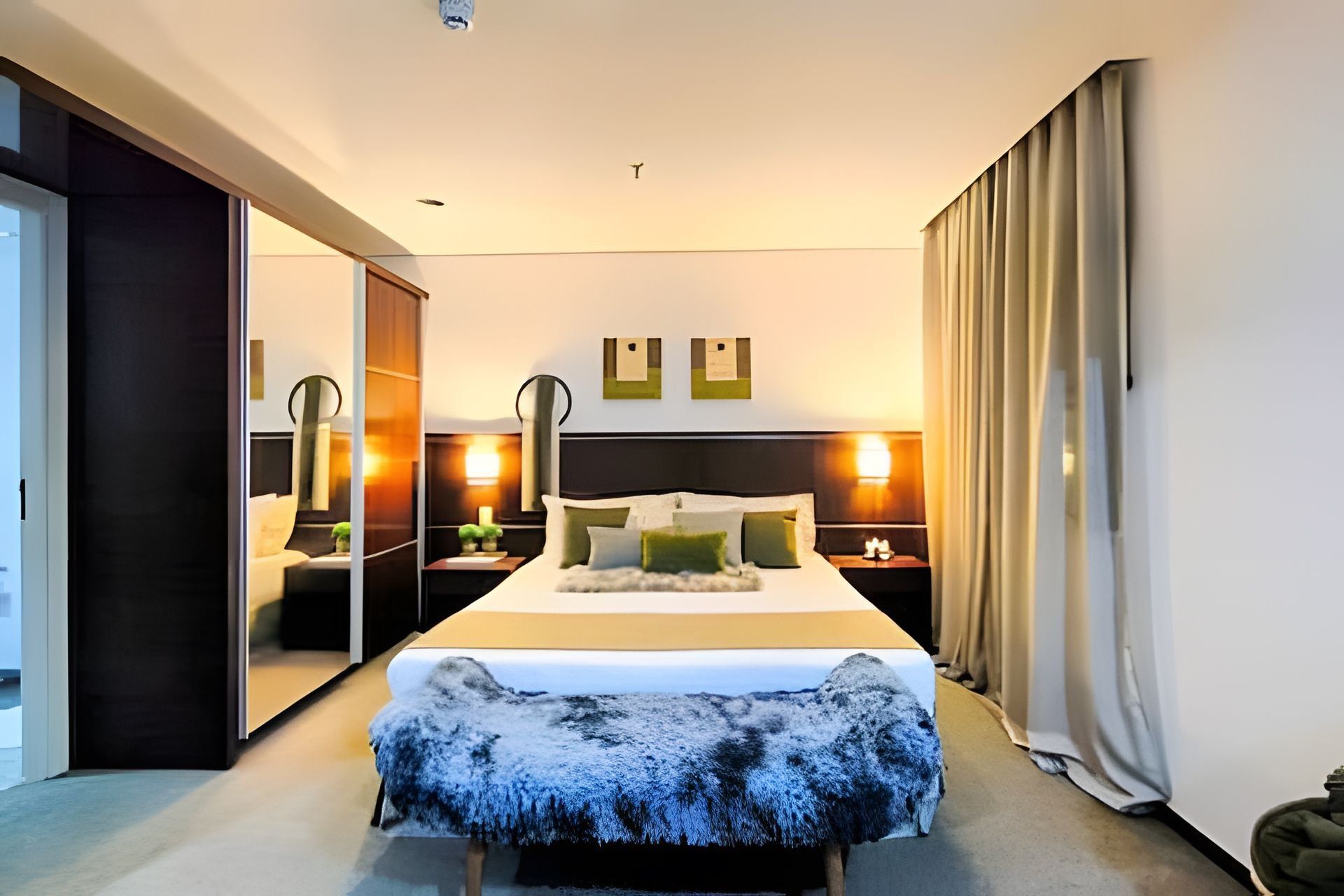 Una habitación de hotel moderna con una cama grande, un espejo, dos mesitas de noche con lámparas, un armario y una puerta corredera.