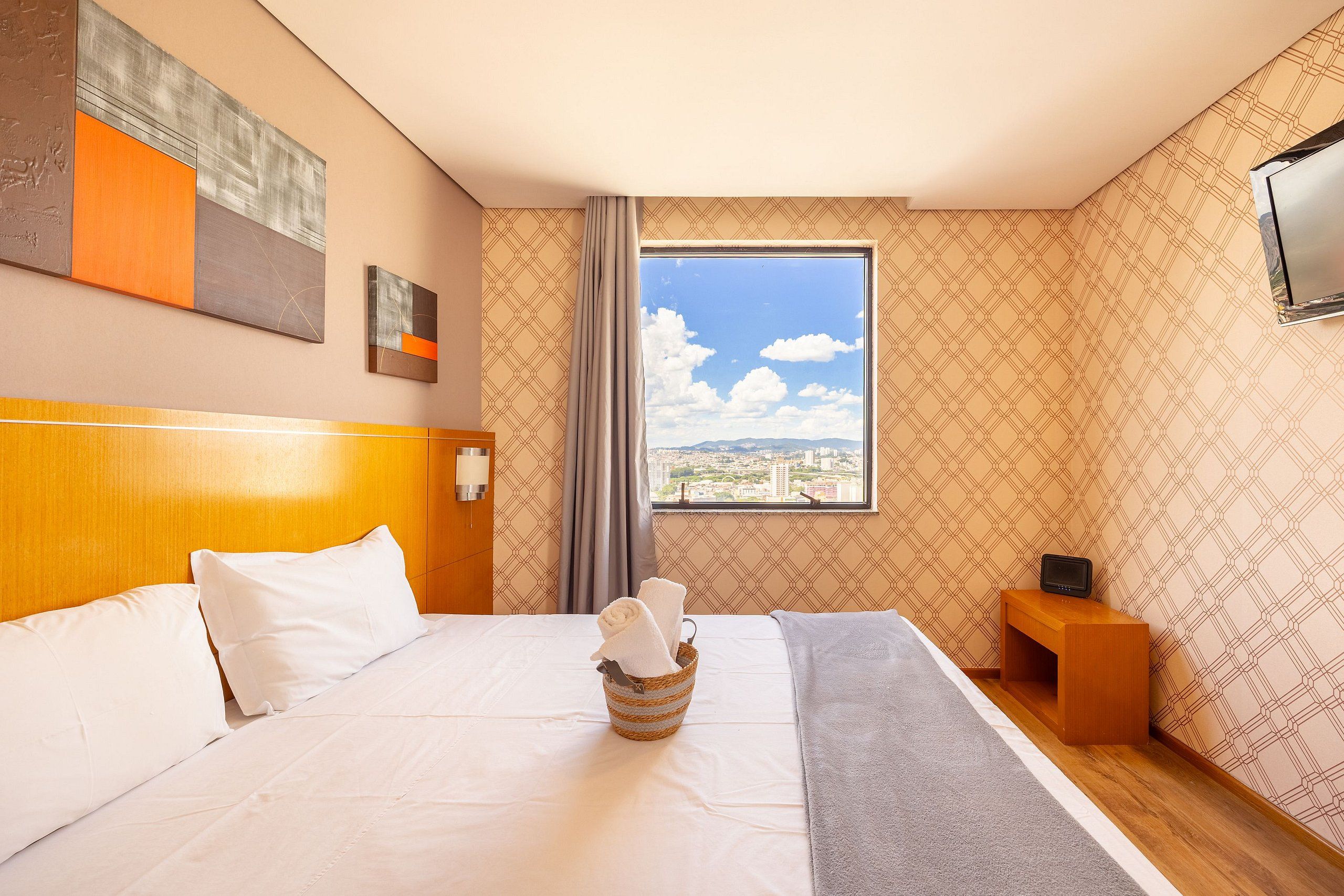 Quarto de hotel com uma cama grande, cabeceira de madeira, janela com vista para a cidade, mesa de cabeceira pequena de madeira e televisão fixada na parede.