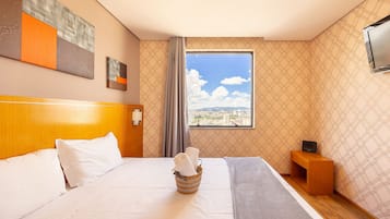 Quarto de hotel com uma cama grande, cabeceira de madeira, janela com vista para a cidade, mesa de cabeceira pequena de madeira e televisão fixada na parede.