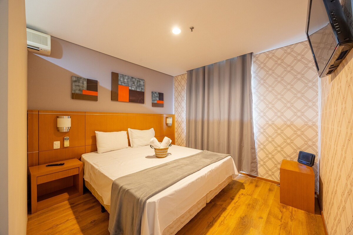 A hotel room with a large bed, a bedside table, a TV, and a wall-mounted air conditioner.