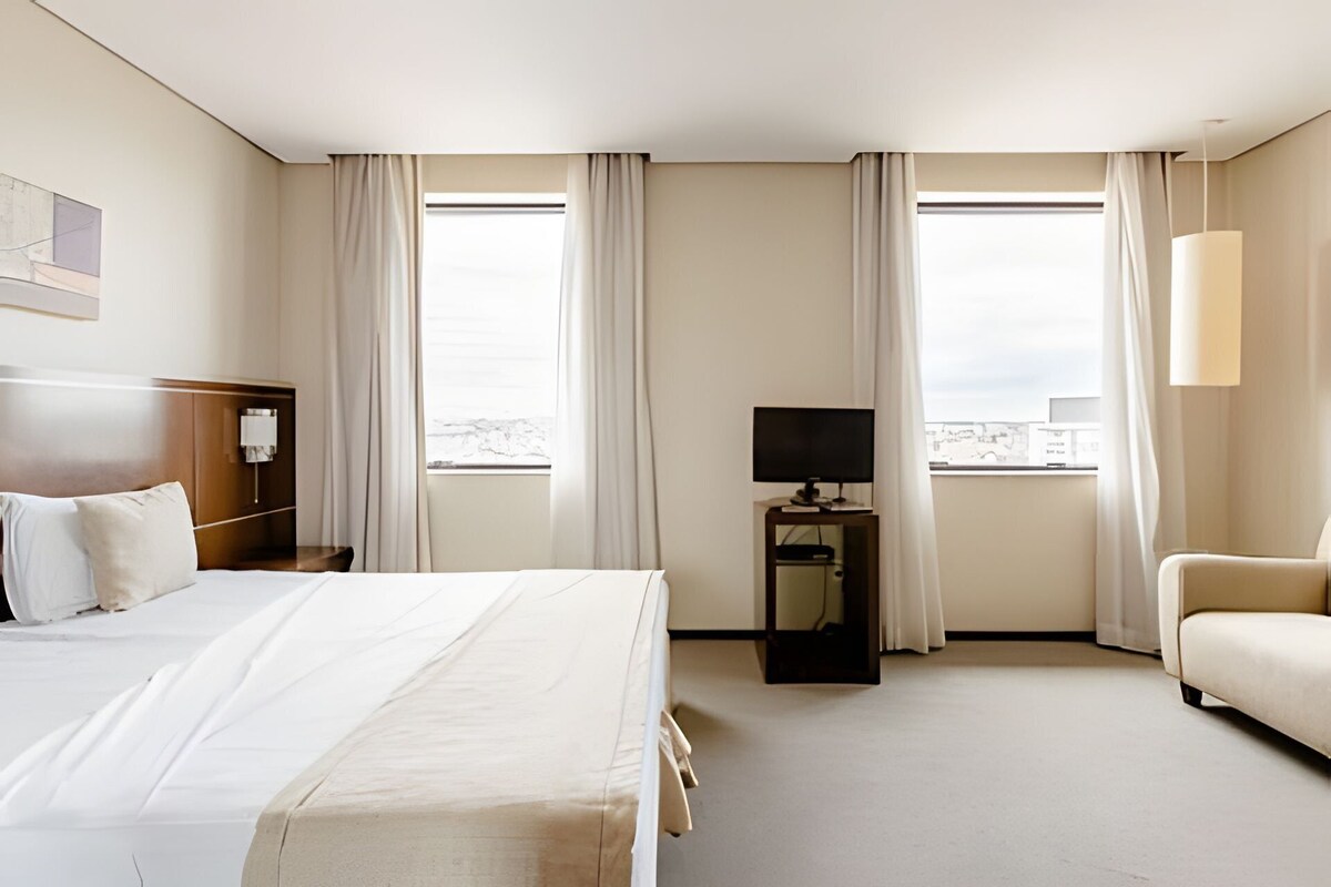 Habitación de hotel con una cama grande, un escritorio con televisor, una silla y dos ventanas con cortinas.