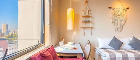 Et soveværelse med en seng, en rød stol, et bord med en lampe, et makramtæppe på væggen og udsigt over byen.