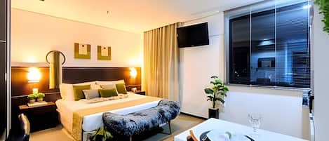 Una habitación de hotel moderna con cama, mesita de noche, lámpara, televisión, ventana y un área de comedor con mesa y sillas.