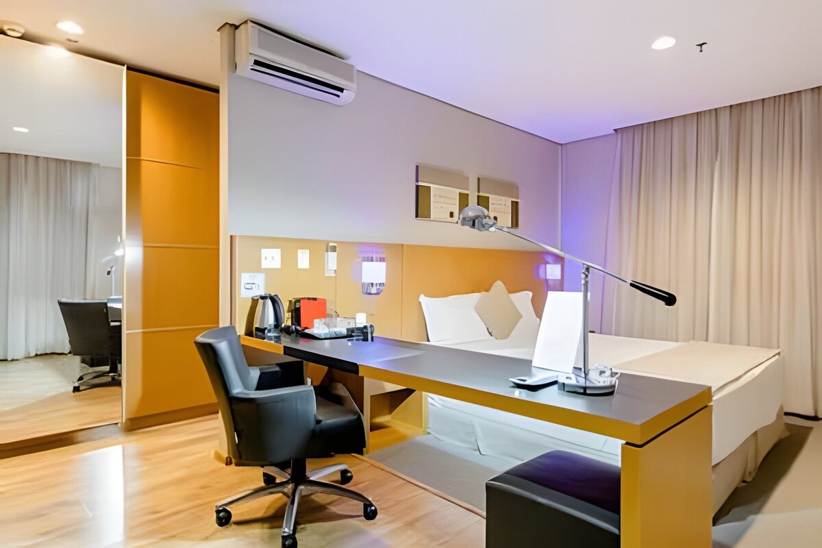 A hotel room with a bed, desk, chair, and an air conditioning unit.