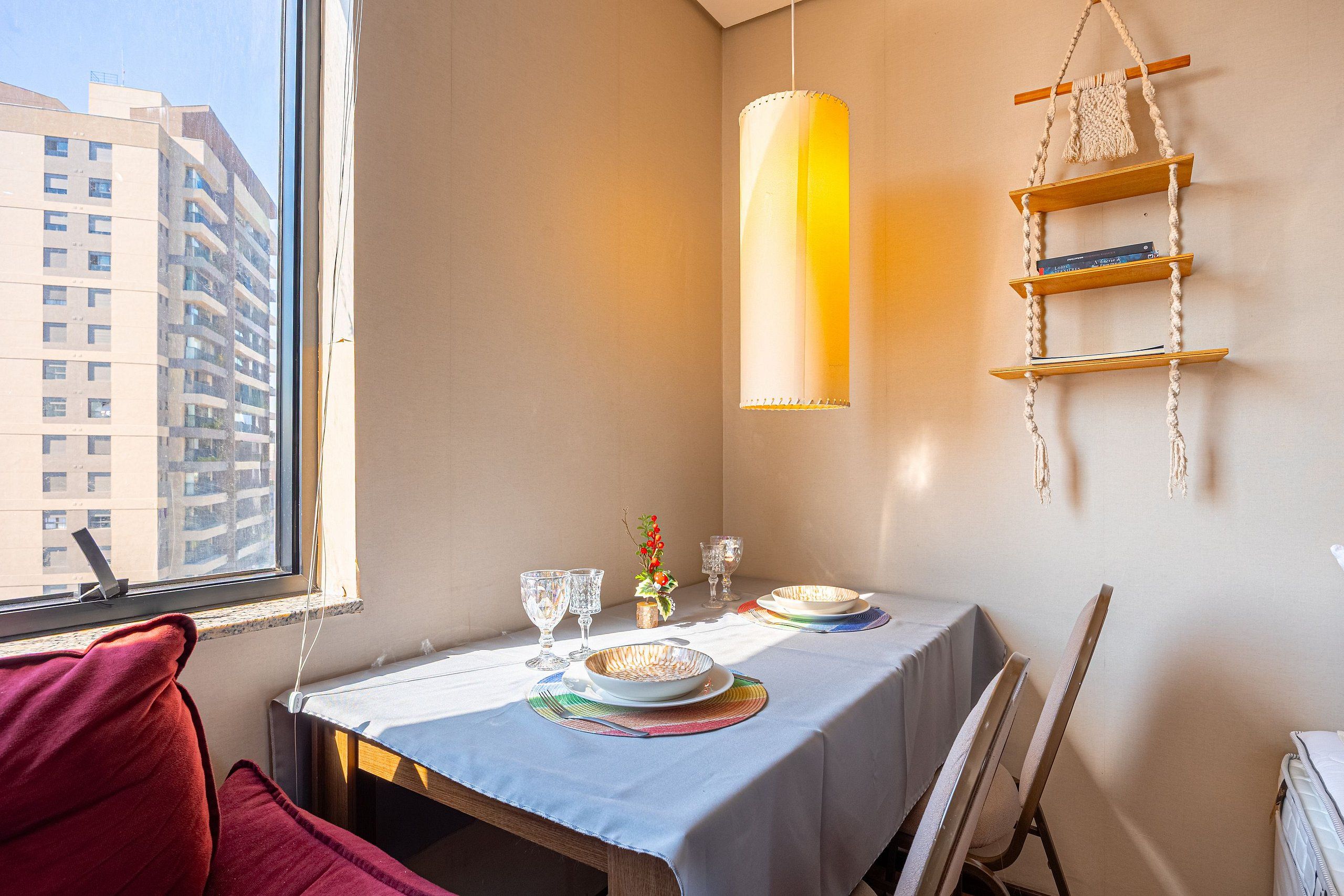 Un área de comedor con una mesa dispuesta para dos, un estante colgante de macramé y vistas a un edificio a través de una ventana.