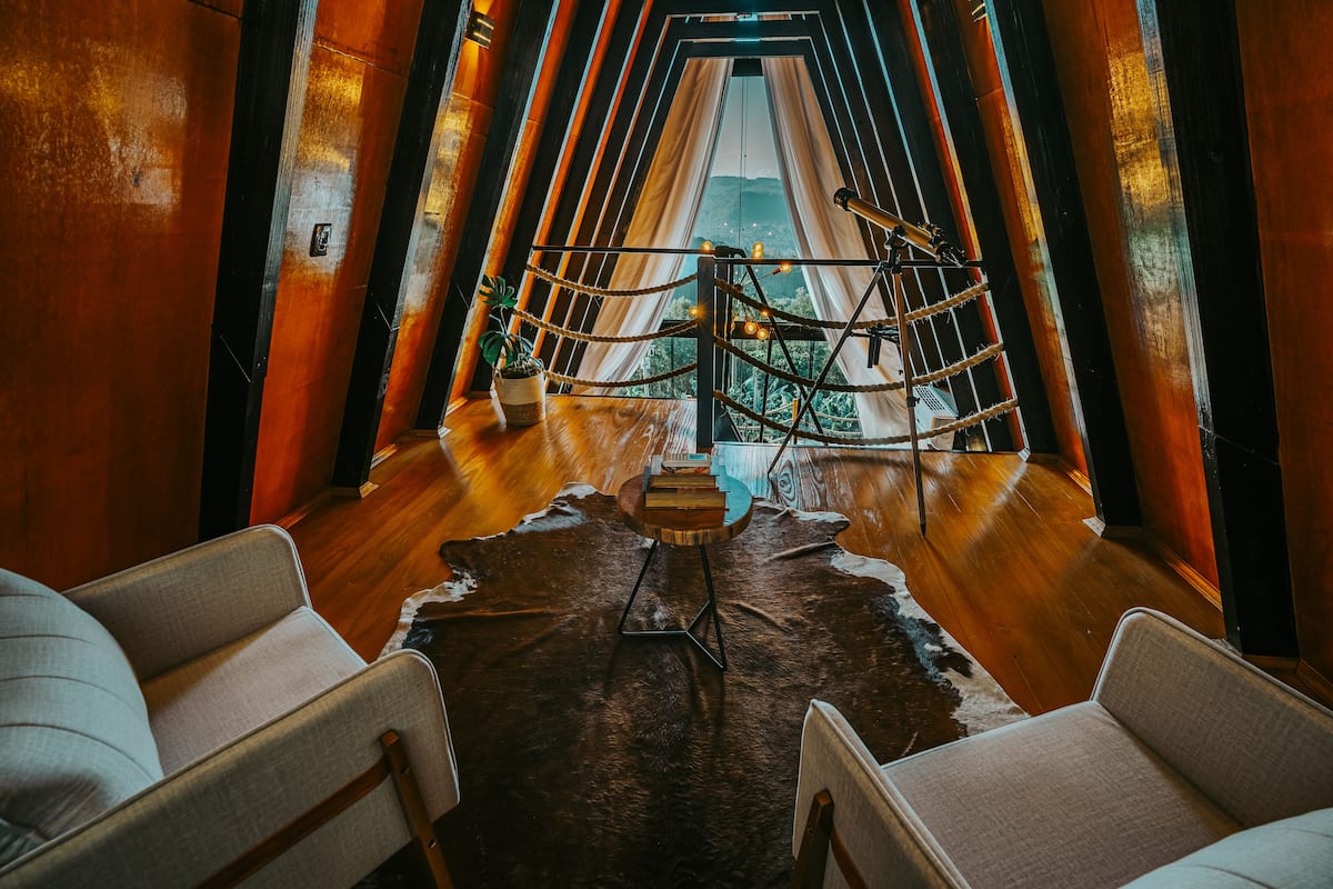 A modern interior with a wooden floor, a central table with books, and a view through a window with curtains.