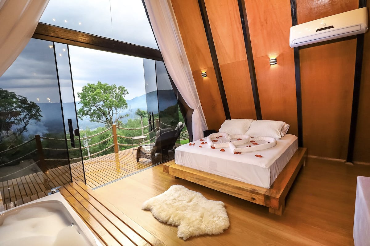 A bedroom with a large window, a wooden bed, a white fur rug, and a view of the outdoors.