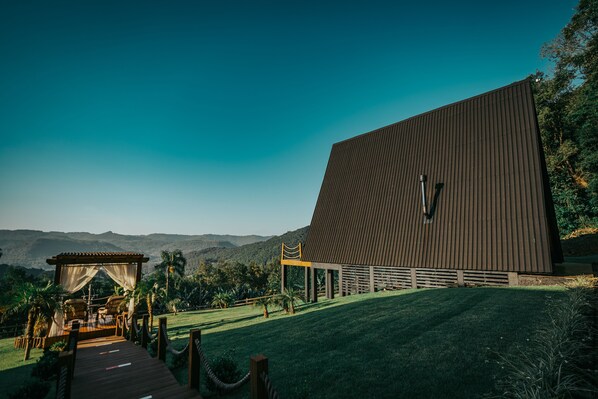 Un bâtiment moderne doté d’un toit en pente, d’une terrasse en bois menant à un espace de détente abrité et offrant une vue sur des collines lointaines.