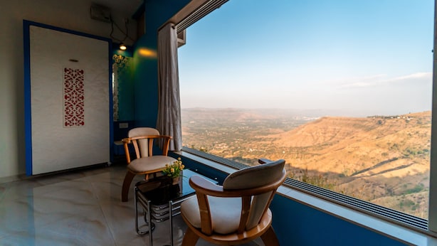 A room with a large window offering a view of a valley. There are two wooden chairs and a small table with a plant. - Treebo Raval Resort with Valley View (Wai)