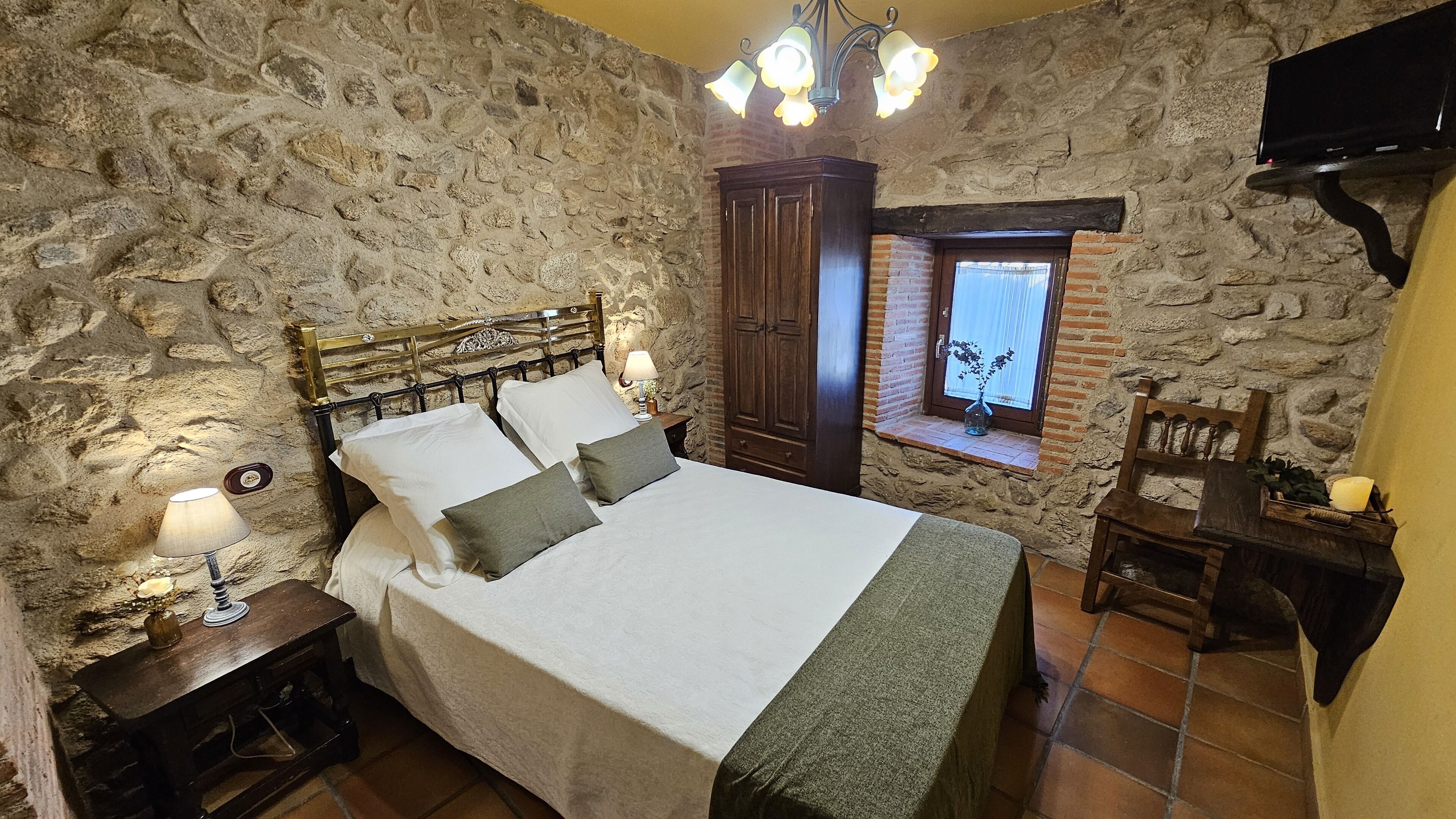 A stone-walled bedroom with a bed, a nightstand, a wooden chair, a window with a plant, and a TV mounted on the wall.
