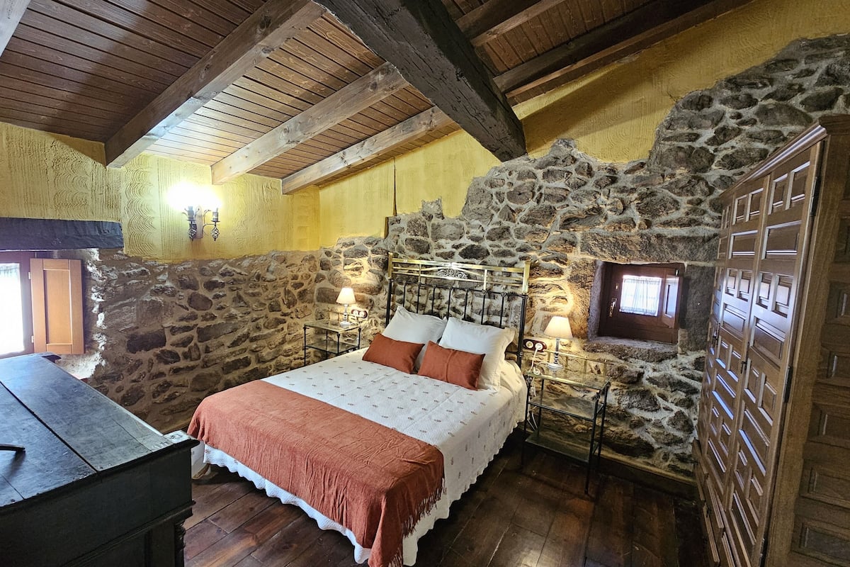 Een landelijke slaapkamer met een stenen muur, houten balken, een bed met een wit en bruin beddegoed en een nachtkastje met een lamp.