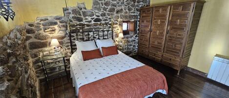 A bedroom with a stone wall, wooden wardrobe, and a bed with a patterned bedspread.