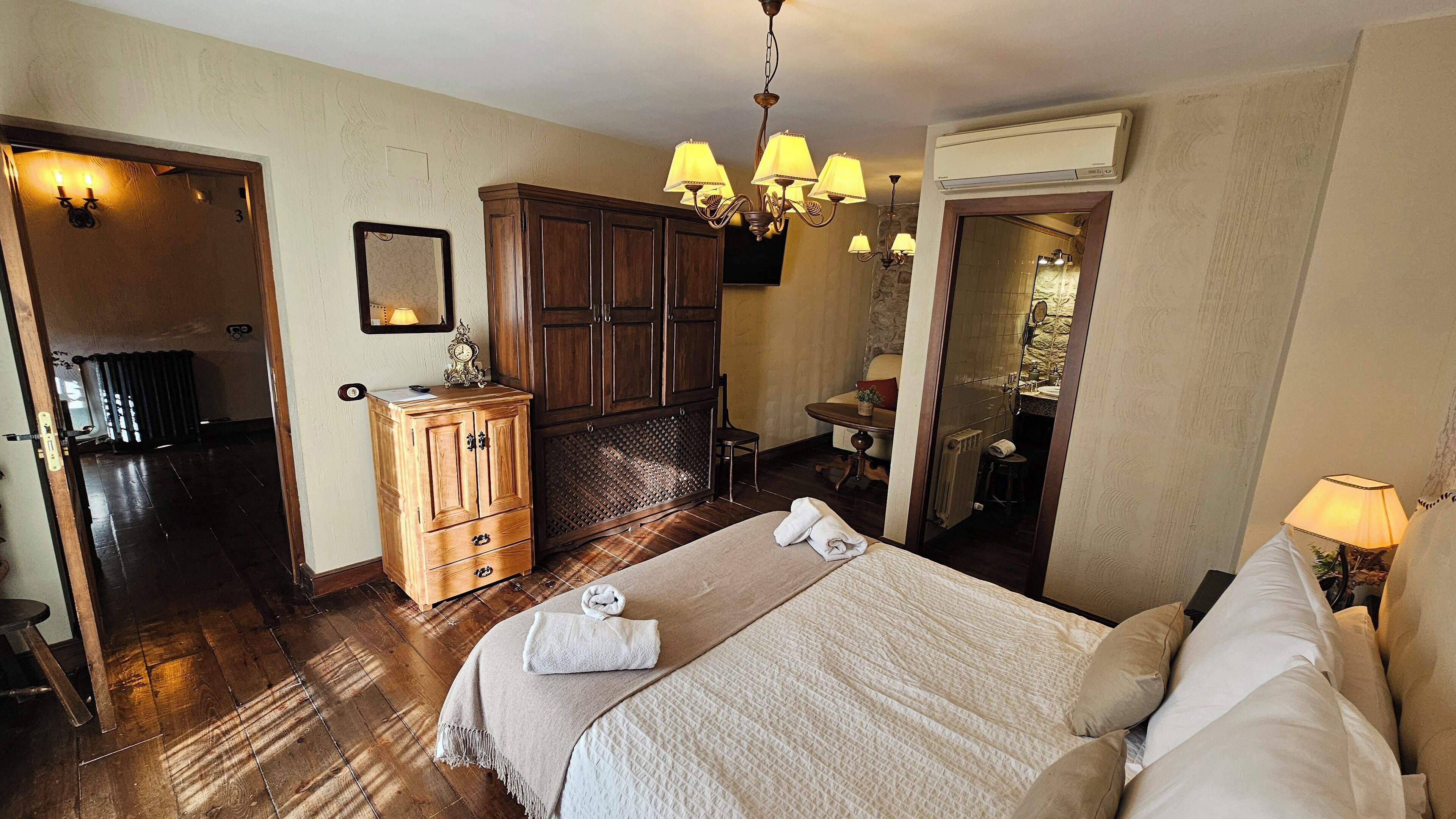 A hotel room with a wooden wardrobe, a bed with a beige blanket, a chandelier, and an air conditioning unit.