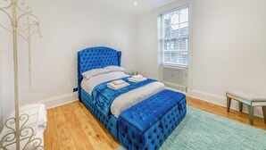 Une chambre avec un lit bleu à capitons, des oreillers blancs, une couette bleue et blanche, un pouf assorti, un radiateur blanc, une fenêtre avec des stores, un présentoir en métal blanc et un plancher en bois.