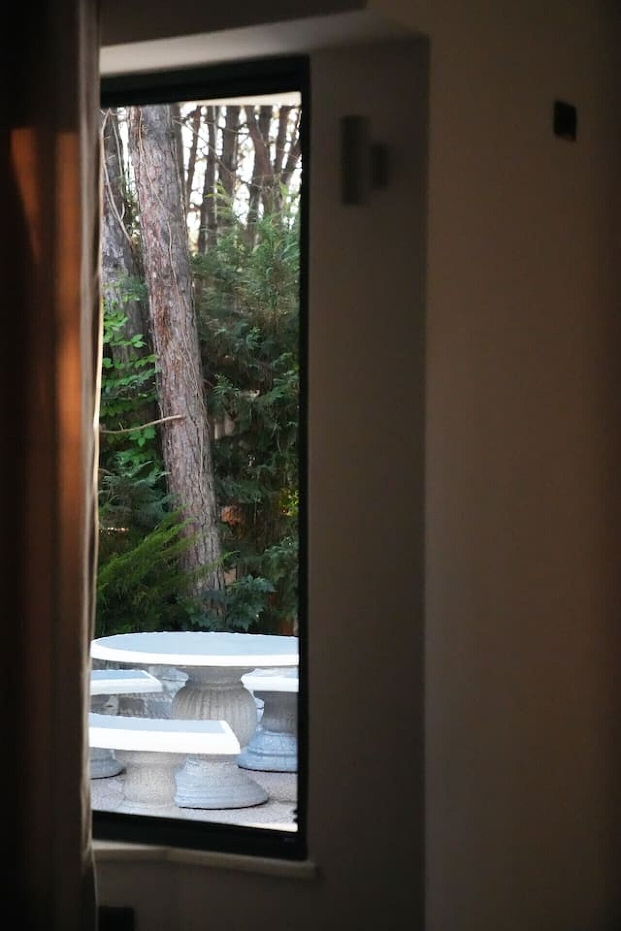 Une vue à travers une fenêtre avec un cadre en bois, un banc en pierre et un arbre à l’extérieur.