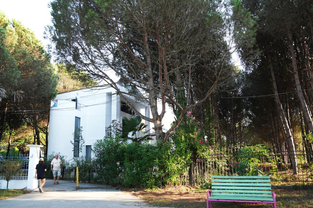 A white building with a large tree in front. There is a green bench and a fence.