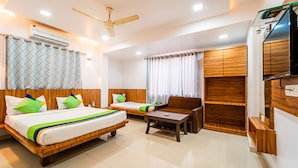 A hotel room with a bed, a sofa, a small table, a TV, and an air conditioning unit. - Itsy Hotels Anupam (Mahabaleshwar)