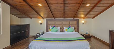 A bedroom with a wooden ceiling, a bed with a white bedspread and colorful pillows, two bedside tables with lamps, and a wooden bench at the foot of the bed.