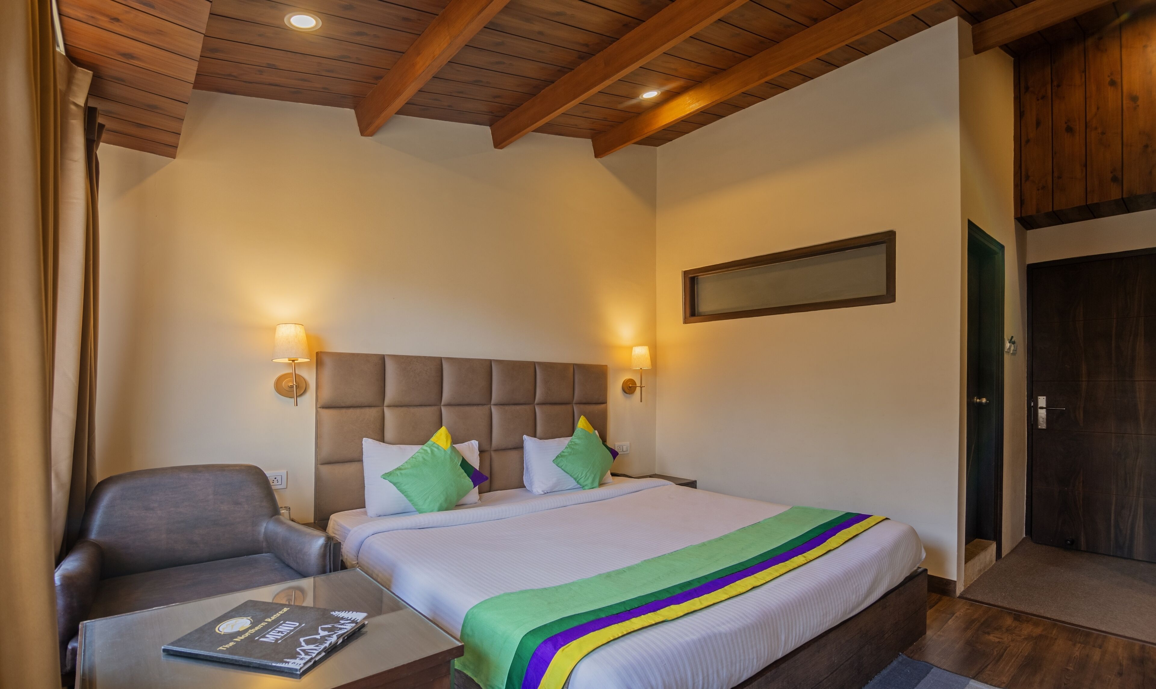 A hotel room with a bed, a chair, a table, and a wall-mounted lamp.