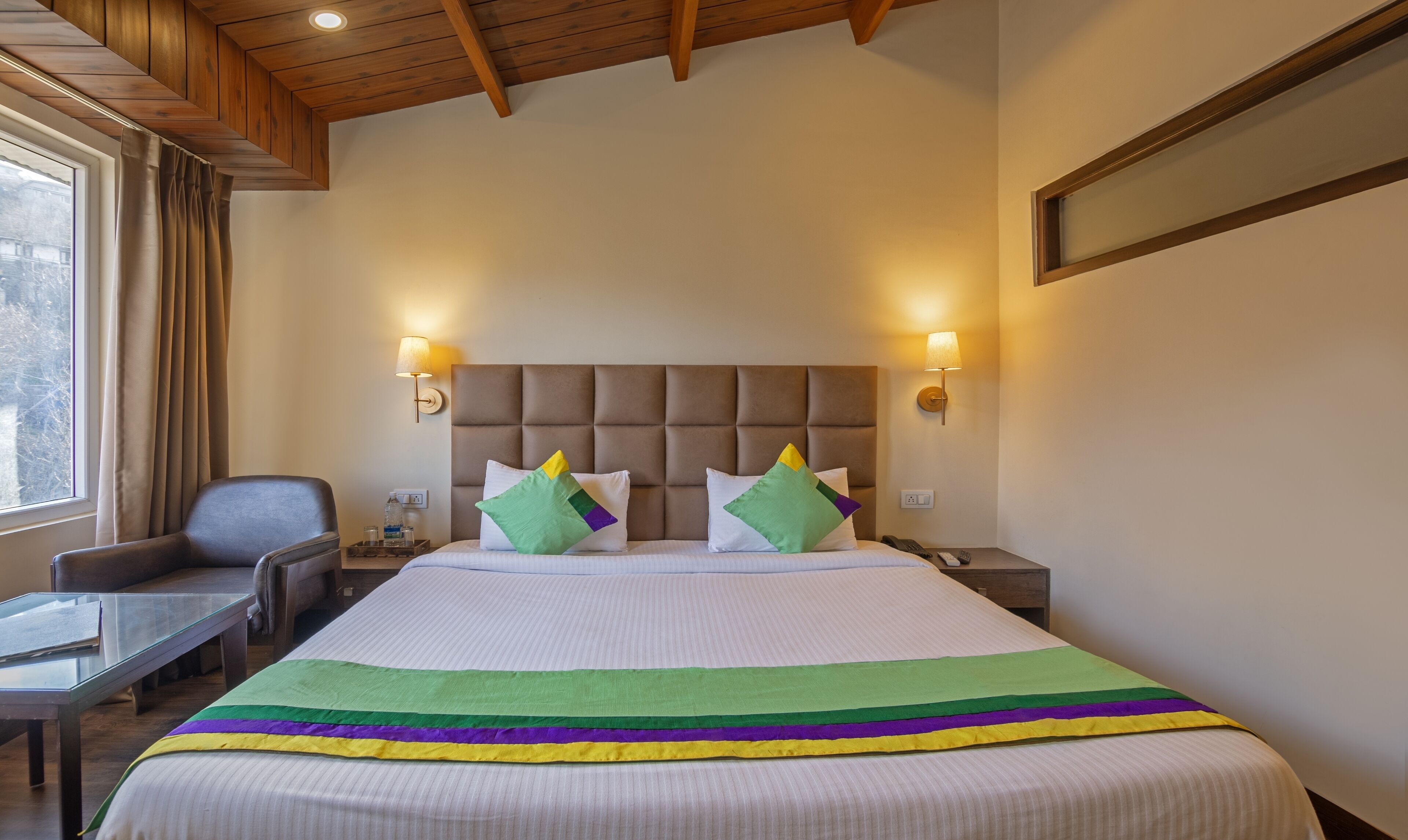 A hotel room with a large bed, a wooden ceiling, two bedside lamps, a chair, a small table, and a window with curtains.