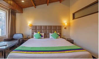 Habitación de hotel con una cama grande, dos lámparas de noche, una silla, una mesita y un ventanal con cortinas.