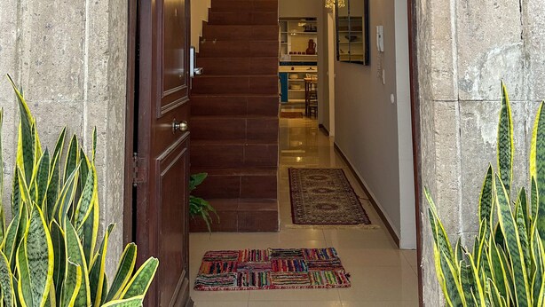 Un pasillo con una escalera de madera que conduce a una plataforma, una araña y una puerta con una cámara de seguridad encima.
