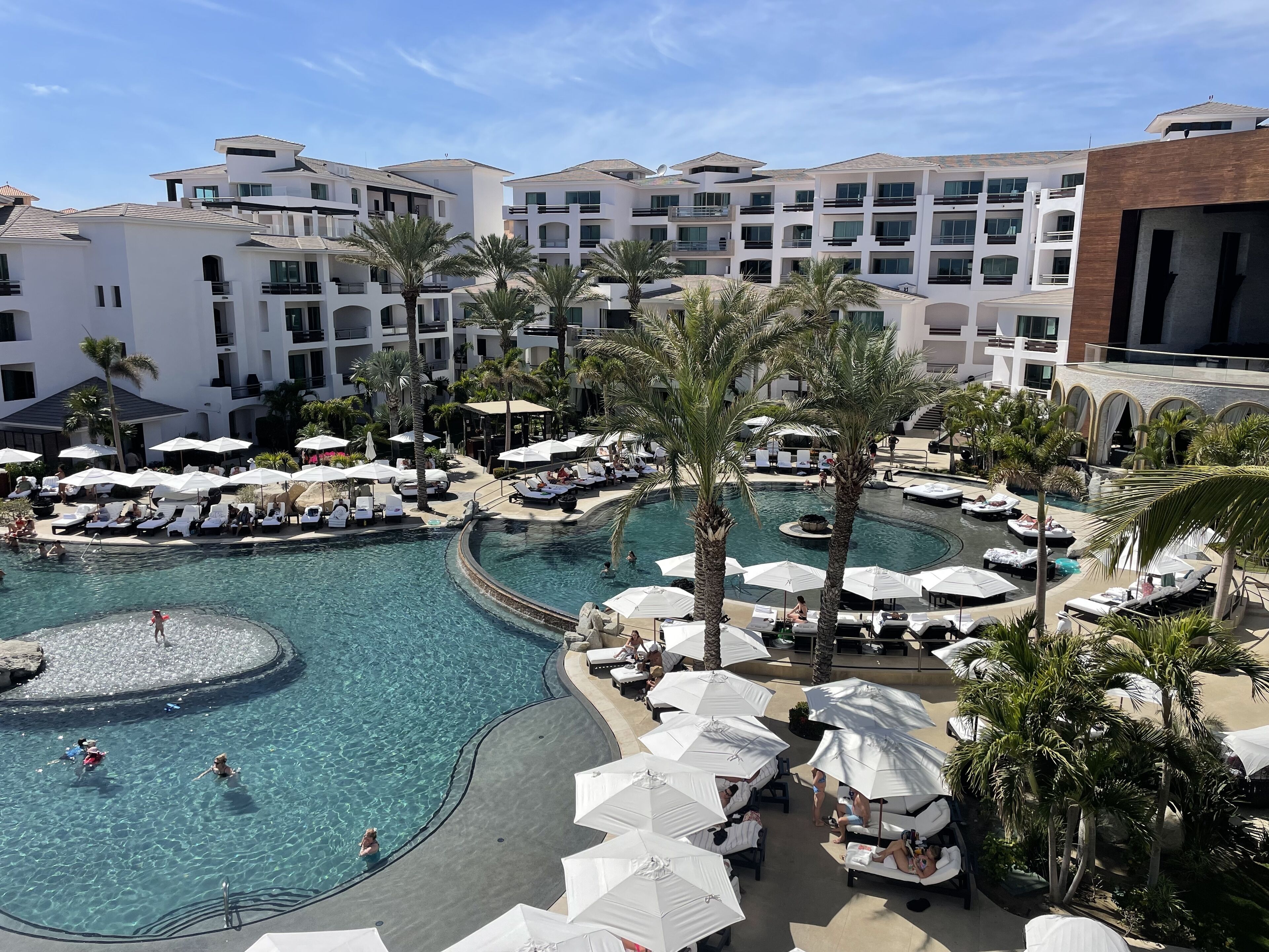 A resort with a central swimming pool, sun loungers, and palm trees.