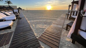 Ett trädäck som leder fram till strandstolar och parasoller, med fri utsikt över solnedgången över havet.