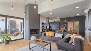 Un salon moderne avec un canapé gris, une table basse et une cheminée. On aperçoit, au fond, des plantes, un coin repas et une cuisine.
