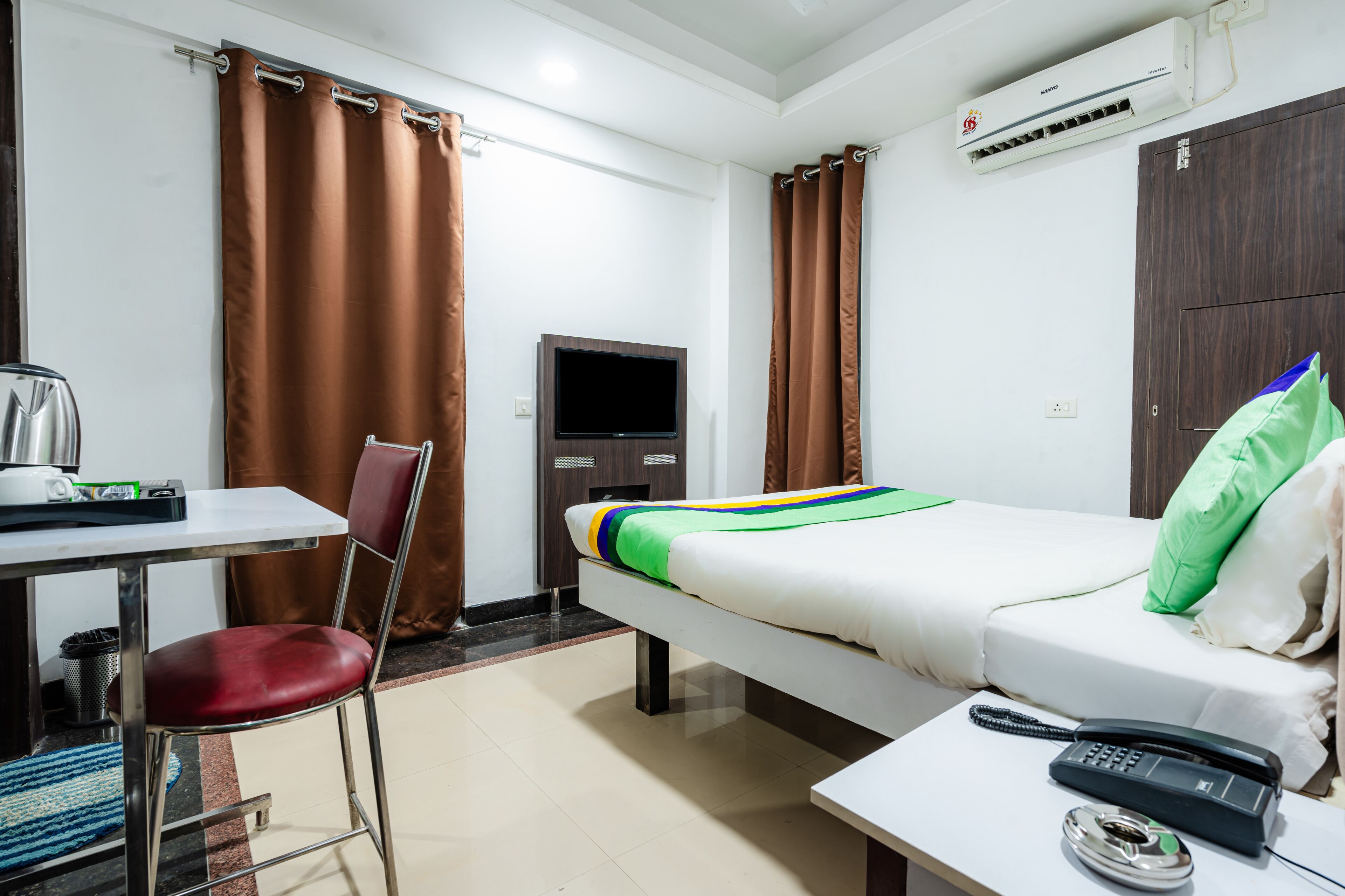 A hotel room with a bed, a desk with a chair, a TV, and an air conditioning unit.