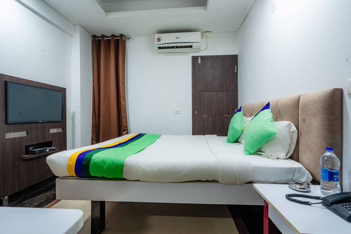 A hotel room with a bed, a TV, an air conditioner, a desk with a phone, and a water bottle.