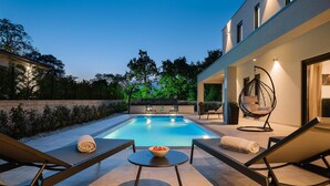 Un espace piscine extérieur moderne, doté de chaises longues, d’un fauteuil suspendu et d’une petite table sur laquelle se trouve un saladier rempli de fruits.