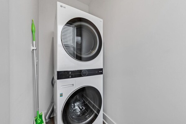Vue de face d’un appareil combiné lave-linge et sèche-linge blanc de la marque LG, fixé au mur, avec une serpillière verte et un tuyau d’eau sur le côté gauche.