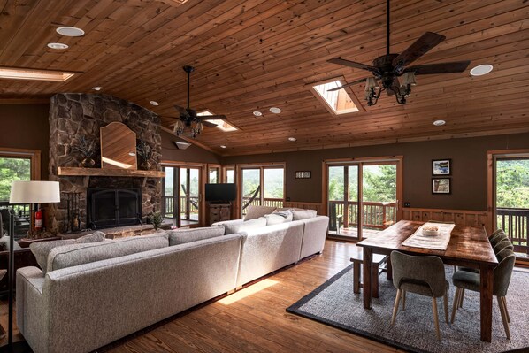 A spacious living area with a stone fireplace, large windows, and a wooden ceiling.
