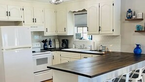 Een moderne keuken met witte kasten, een groot eiland met barstoelen, een zwarte prullenbak en een raam met blinds.