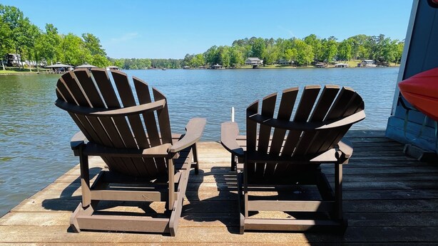 Twee Adirondack stoelen op een houten steiger, met een meer en huizen op de achtergrond.