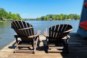 Dos sillas Adirondack sobre un muelle de madera, con un lago y casas al fondo.