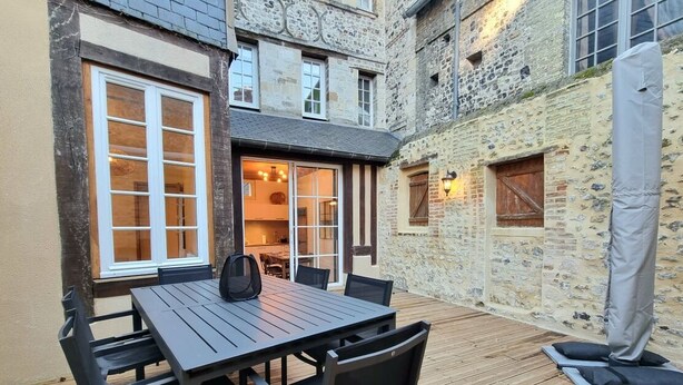 A courtyard with a wooden table and chairs, a covered seating area, and a stone wall with windows.