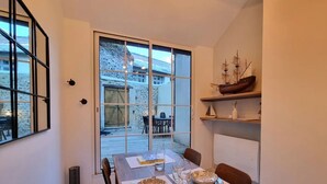 A dining area with a table set for two, a large window with a view of a courtyard, and a ship model on a shelf.