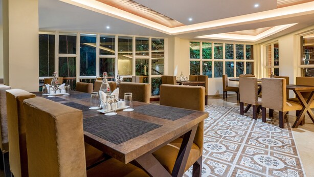 A dining area with multiple tables and chairs, large windows with views of greenery, and a patterned floor. - Itsy Hotels Meraki Grand (Gangtok)