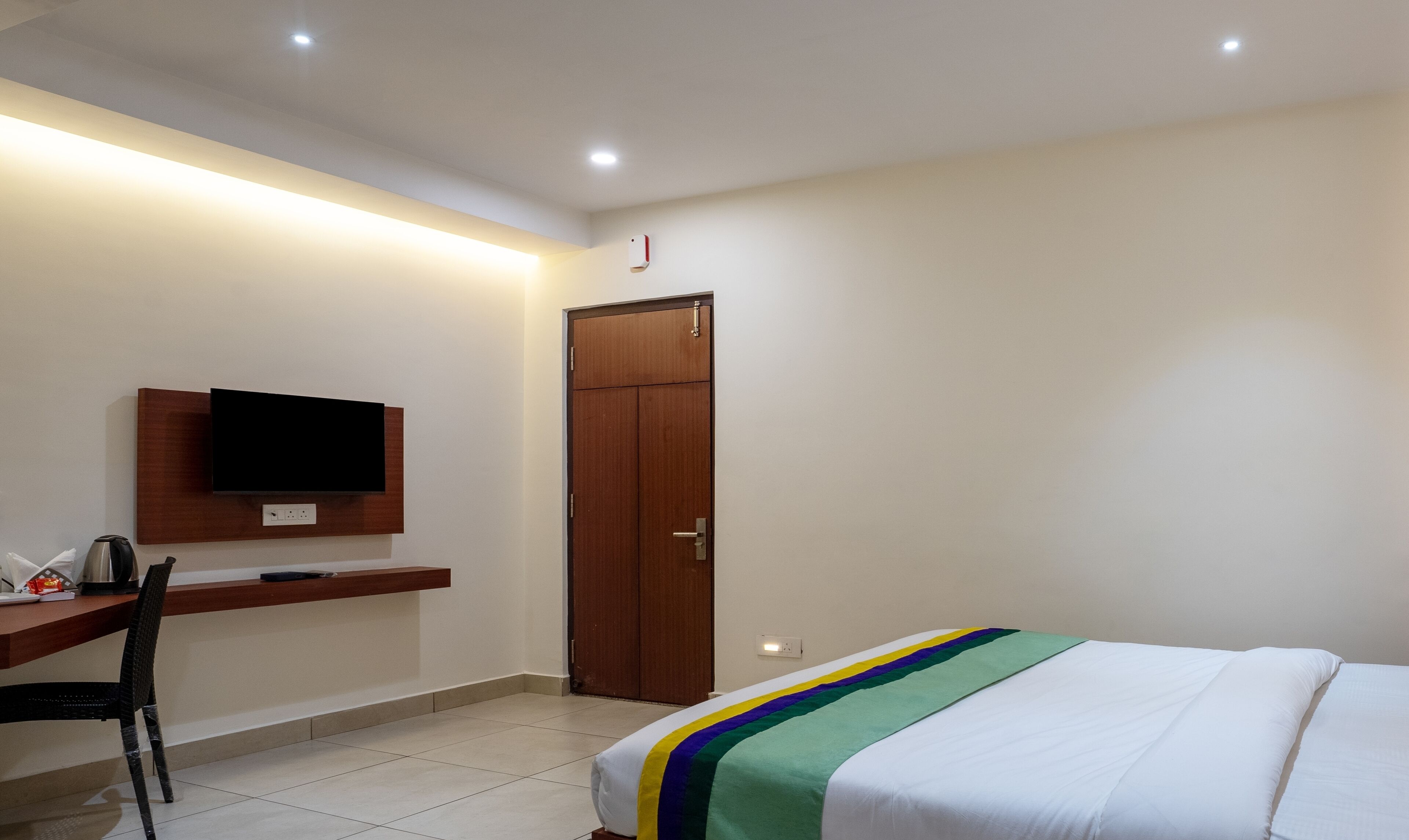 A hotel room with a bed, a desk with a chair, a television mounted on the wall, and a wooden door.