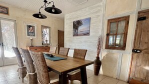 A dining area with a wooden table and wicker chairs, a vase with dried plants, and a beach scene painting on the wall.