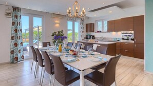 A modern kitchen with wooden cabinets, a dining area with a white table set for six, and a view of greenery outside.