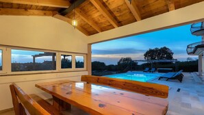 A wooden dining table with benches, a pool, and a view of the surrounding landscape through large windows.