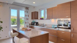 A modern kitchen with wooden cabinets, a granite countertop, and a built-in oven. There are two white bar stools and a window with a view of the outdoors.