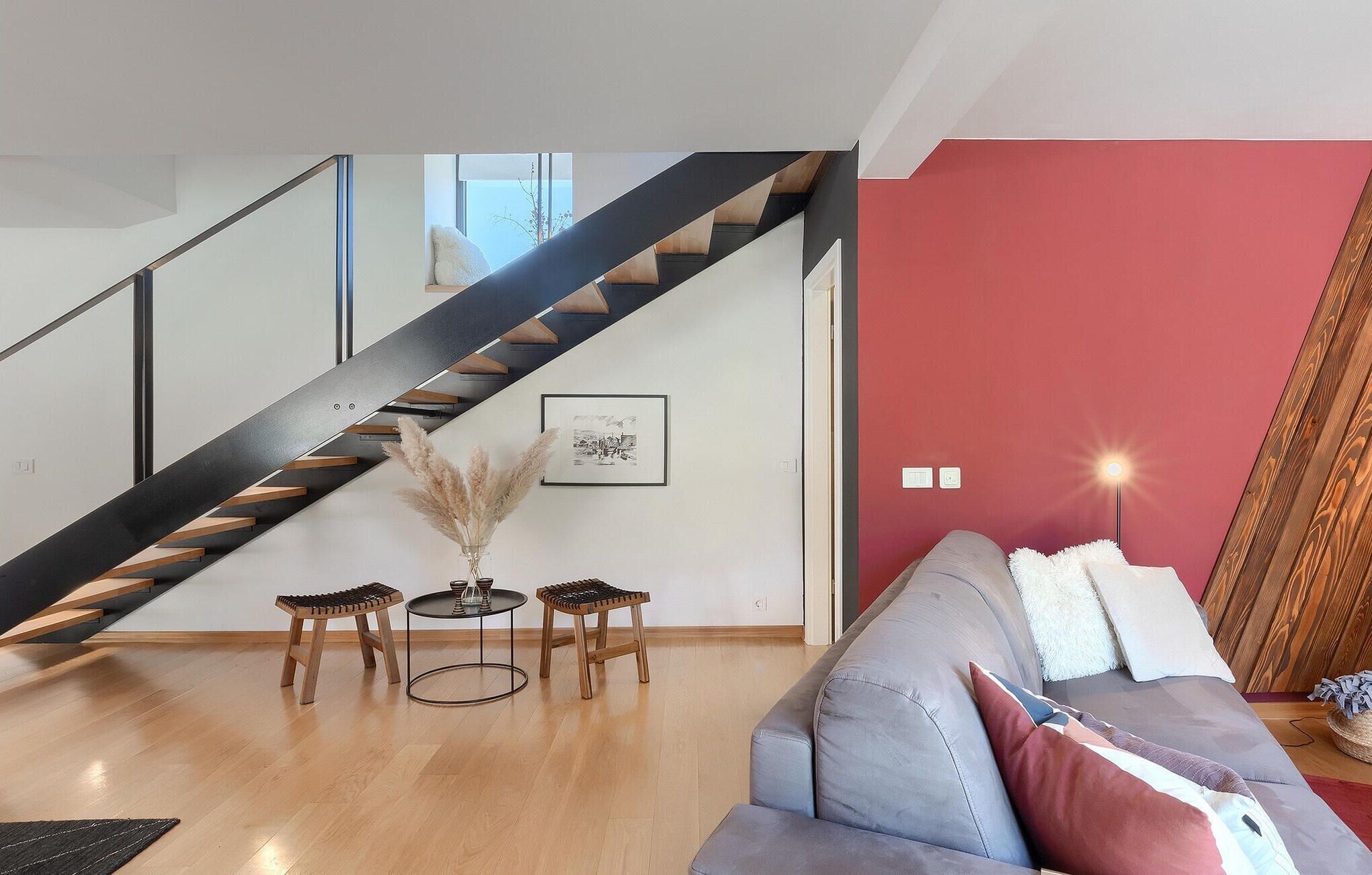 A modern living room with a staircase, a gray sofa, and a red accent wall.