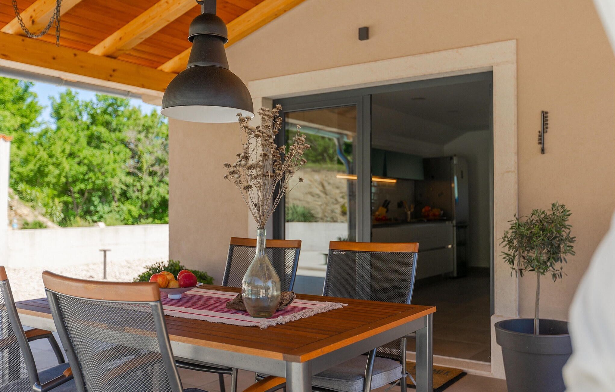 An outdoor dining area with a wooden table, metal chairs, a hanging pendant light, and a vase with dried flowers.
