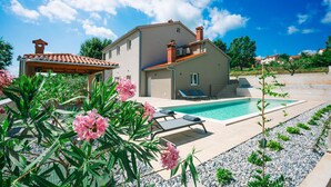 Una casa a due piani con piscina, area relax esterna e giardino fiorito di fiori rosa.