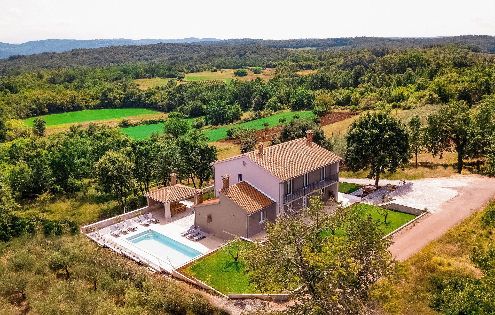 Una casa de dos pisos con piscina, rodeada de abundante vegetación y campos.