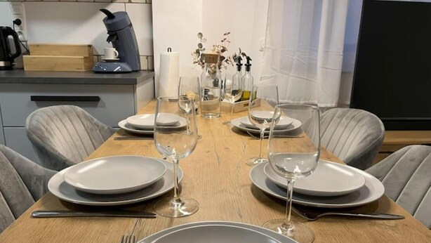 Een eettafel gedekt met borden, glazen en bestek, met een koffiezetapparaat en decoratieve items op het aanrecht.